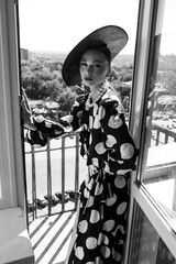 Woman with black hat and polka dot dress posing for fashion on a balcony with fresh air and sun - 396099019