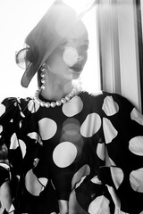 Woman with black hat and polka dot dress posing for fashion on a balcony with fresh air and sun - 396099023