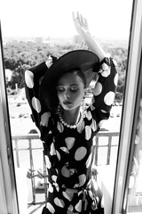 Woman with black hat and polka dot dress posing for fashion on a balcony with fresh air and sun - 396099053