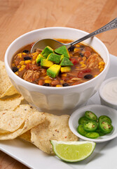 Bowl of chicken taco soup with tortilla chips in a white bowl garnished with avocado with sides of Serrano chili pepper and sour cream; copy space