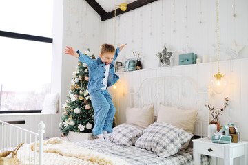 little baby boy in pajamas is happy and jumping on the bed on the background of Christmas tree and gifts.