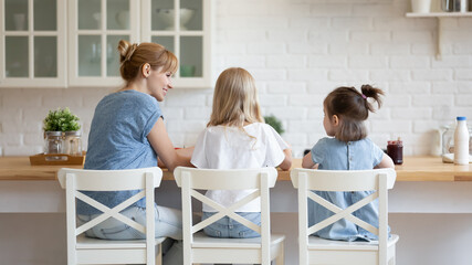 Rear view smiling young mother with two little daughters sitting at modern kitchen table, family enjoying breakfast together, adorable preschool girls enjoying weekend, preparing dinner