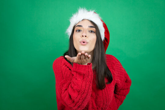Young beautiful woman wearing Christmas Santa hat over isolated green background looking at the camera blowing a kiss with hand on air being lovely and sexy. Love expression.