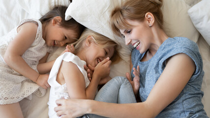 Top view overjoyed mother and two little daughters lying in cozy bed together, hugging and...