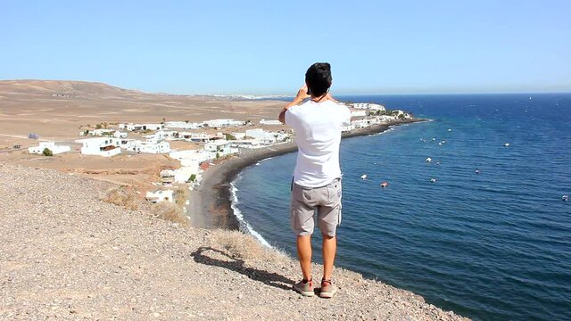 Young man walks to cliff edge to take photo of Puerto Calero coast town by the ocean. Male tourist using cellphone to capture beautiful natural landscape in Lanzarote island, Spain