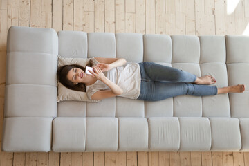 Top view happy beautiful young woman using phone, relaxing on couch at home, smiling female lying...