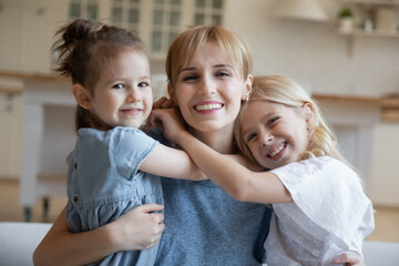 Head shot portrait smiling young mother hugging two little daughters, looking at camera, overjoyed beautiful mum and adorable kids cuddling, posing for family photo at home, having fun