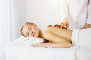 Obraz na płótnie Canvas Beautiful caucasian woman enjoying warm stones procedure in sunny spa salon. Beauty concept