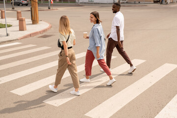 Young happy intercultural friends having drinks and talking while crossing road