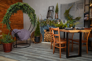 Rustic style Christmas festive decorations, fireplace with firewood, table, chairs, circle sitting swing with fresh fir branches, grey thick-yarn knitted plaid on seat. Eco-friendly decorative element