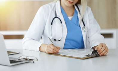 Unknown woman-doctor at work filling up medication history record form in sunny clinic, close-up