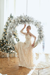 Caucasian woman with hairstyle in white pink long dress posing in room with large windows and full of sun and bright and Christmas tree on a wooden floor - 396116427