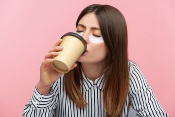 Tired sleepy woman with brown hair in striped shirt doing beauty procedure wearing eye patches and...