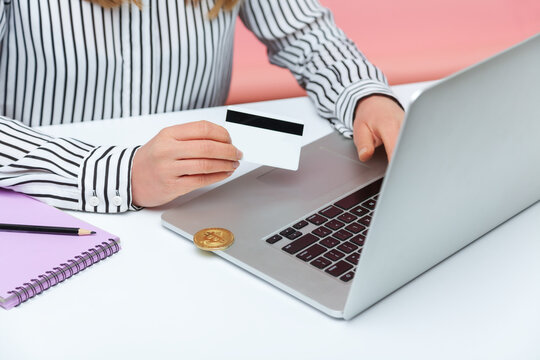 Closeup female holding credit card typing on laptop, bitcoin crypto coin lying on pc corner, withdrawal of electronic money to the card, banking. Indoor studio shot isolated on pink background