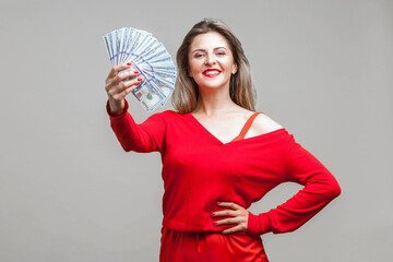 Portrait of arrogant wealthy beautiful woman in red dress standing showing dollar bills, looking at camera with smile, proud of big money, income. indoor studio shot isolated on gray background