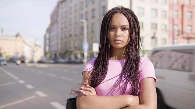 A young black woman looks seriously at the camera in the street in an urban area - a busy road in the blurry background