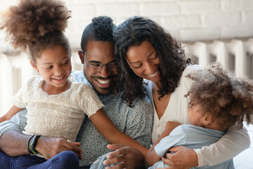 Strong bond. Loving joyful african american millennial spouses husband and wife enjoying relax rest on couch cuddling their children little boy and school age girl playing laughing tickling having fun