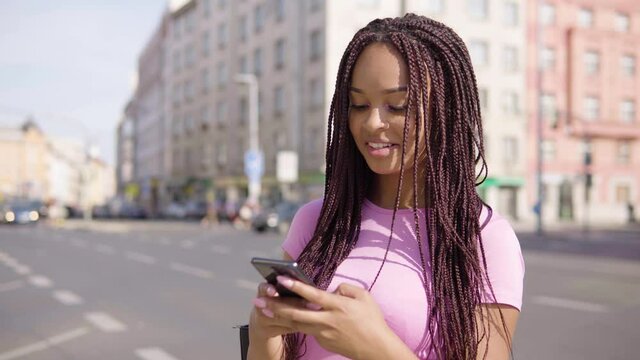 A young black woman works on a smartphone in the street in an urban area - a busy road in the blurry background