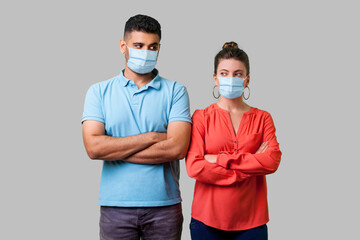 Fototapeta na wymiar Portrait of upset couple with surgical medical mask standing together with crossed hands, looking sideways at each other with resentful glance, suspicion. isolated on gray background, indoor studio