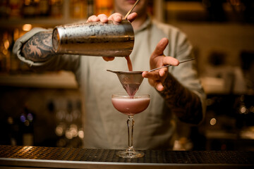 bartender holds sieve over wine glass and pours pink cocktail from shaker.
