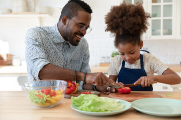 Assisting daddy. Happy smiling single african black dad and small tween daughter engaged in easy cooking together spending time at kitchen talking preparing healthy vegan lunch of lettuce and pepper