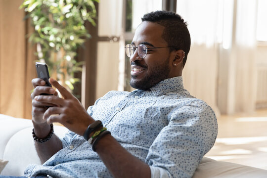 Pleasant leisure time. Peaceful smiling young african american man enjoy relaxing on cozy sofa with gadget, making video call using mobile phone webcam, playing online game by wifi, browsing internet
