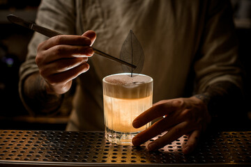 man bartender holds tweezers with blue leaf to decorate glass with cocktail