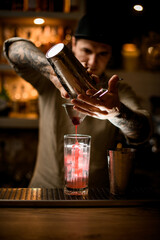 man in hat holds sieve over transparent glass and gently pours cocktail from shaker.