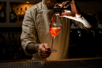 male bartender holds with hand wine glass with bright drink and adds liquid from jigger