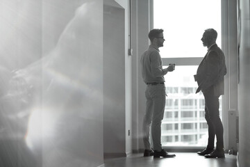 Black and white photo of two businessmen standing in the modern office against panoramic window and discussing something while having coffee break