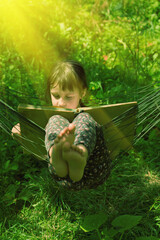 Portrait of young beautiful girl reading the book in  hammock outdoors at green garden. Vertical image.