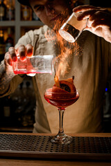 man bartender accurate add ingredients to glass with vegetables inside and makes fire over it
