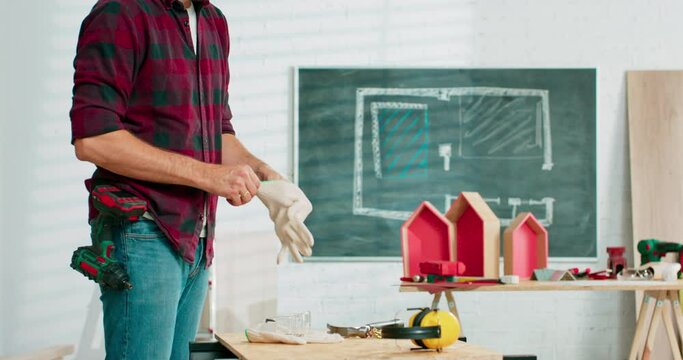 A young carpenter wearing jeans and a plaid flannel shirt puts on white cloth protective gloves before starting work in the carpentry shop. There are a locksmith table and tools in the background.
