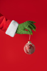 A green hairy hand in a Santa suit holds a red Christmas ball on a red background