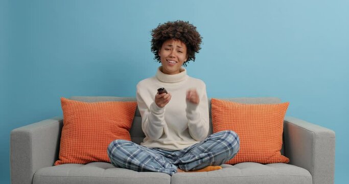 Pleased woman relaxes at home on sofa watches TV switches channels with remote controller enjoys television program poses in modern apartment isolated on blue background has fun. Leisure and people