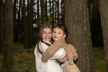 Fototapeta premium A teenage girl in a light dress hugs her little brother. Children are happy and laugh among the trees in forest. Real childhood and family happiness in the middle of nature. Close-up photo