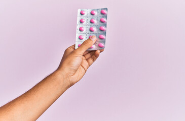 Hand of hispanic man holding pills over isolated pink background.