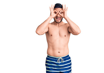 Fototapeta premium Young handsome man wearing swimwear doing ok gesture like binoculars sticking tongue out, eyes looking through fingers. crazy expression.
