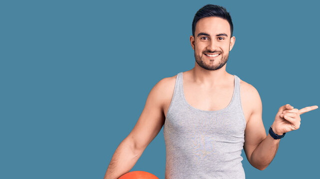 Young handsome man holding basketball ball smiling happy pointing with hand and finger to the side
