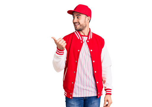 Young handsome man wearing baseball jacket and cap smiling with happy face looking and pointing to the side with thumb up.