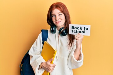 Young red head girl wearing backpack holding back to school banner clueless and confused expression. doubt concept.