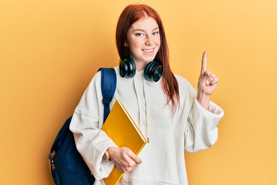 Young red head girl wearing student backpack and holding book smiling with an idea or question pointing finger with happy face, number one