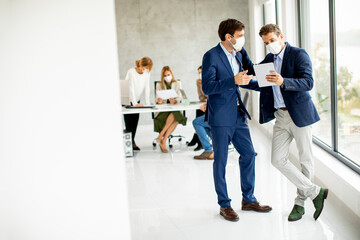 Two young business men with facial protective masks discussing  with paper plan in the office