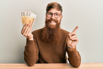 Young irish redhead man holding 500 norwegian krone banknotes sitting on the table smiling happy...