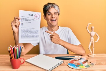 Young hispanic man artist holding passed exam sitting on the table smiling happy pointing with hand and finger