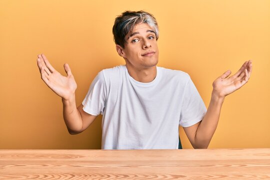 Young hispanic man wearing casual clothes sitting on the table clueless and confused with open arms, no idea and doubtful face.