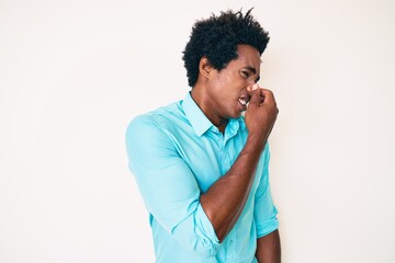 Handsome african american man with afro hair wearing casual clothes smelling something stinky and disgusting, intolerable smell, holding breath with fingers on nose. bad smell