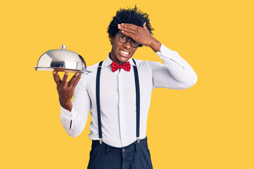 Handsome african american man with afro hair wearing waiter uniform holding silver tray stressed and frustrated with hand on head, surprised and angry face