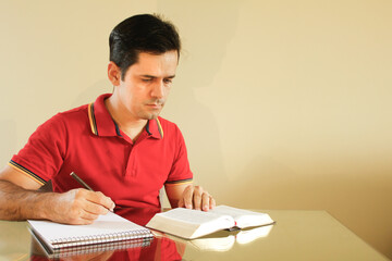Adult man sitting at the table in his living room, with yellow background wall, reading the bible...