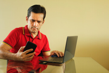 Adult man sitting at desk in living room of his house, with yellow background wall, operating his notebook and looking at his smartphone